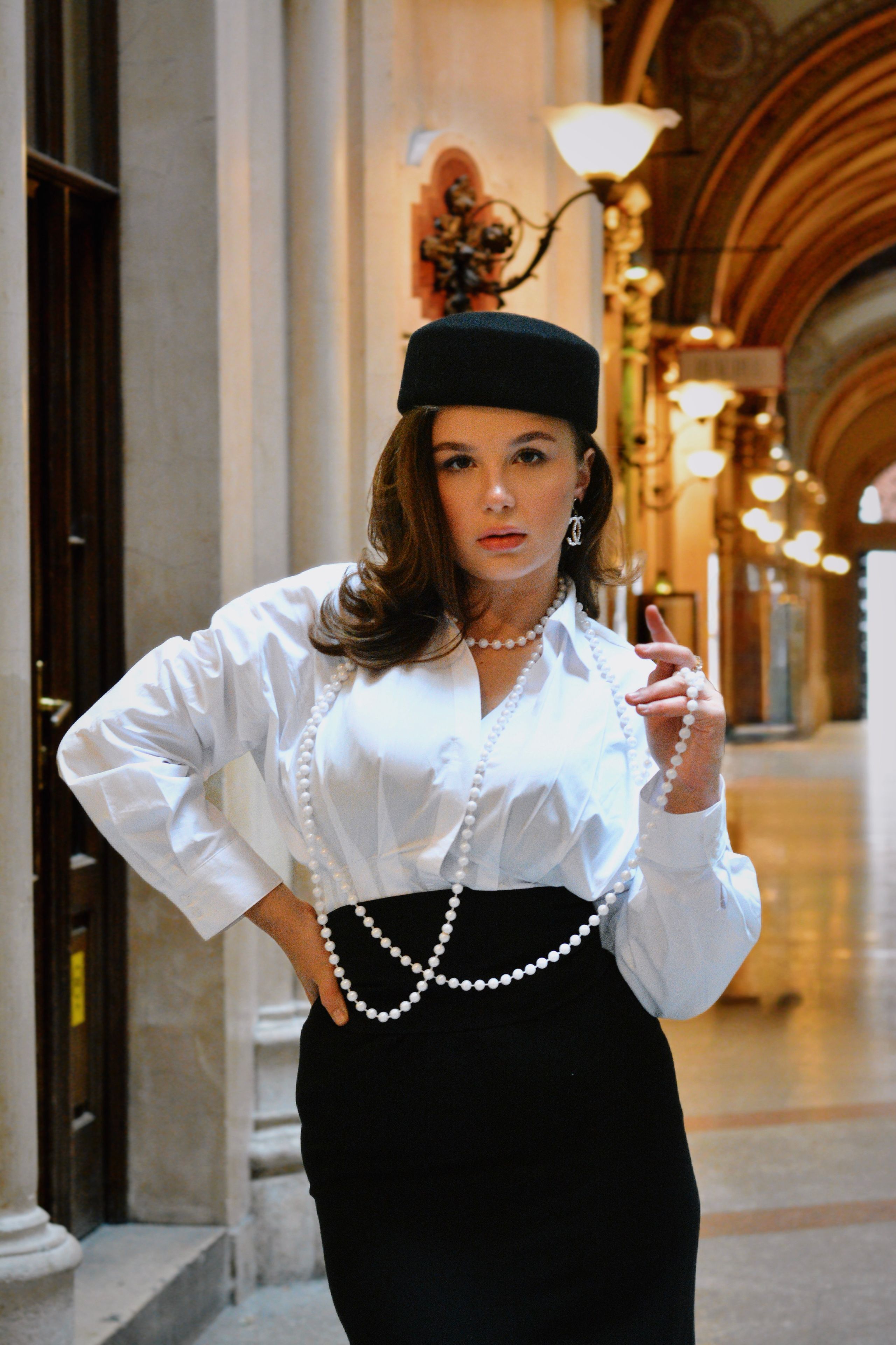A stylish woman in a white blouse and black skirt poses in a grand, classical Viennese hallway, capturing an elegant portrait.