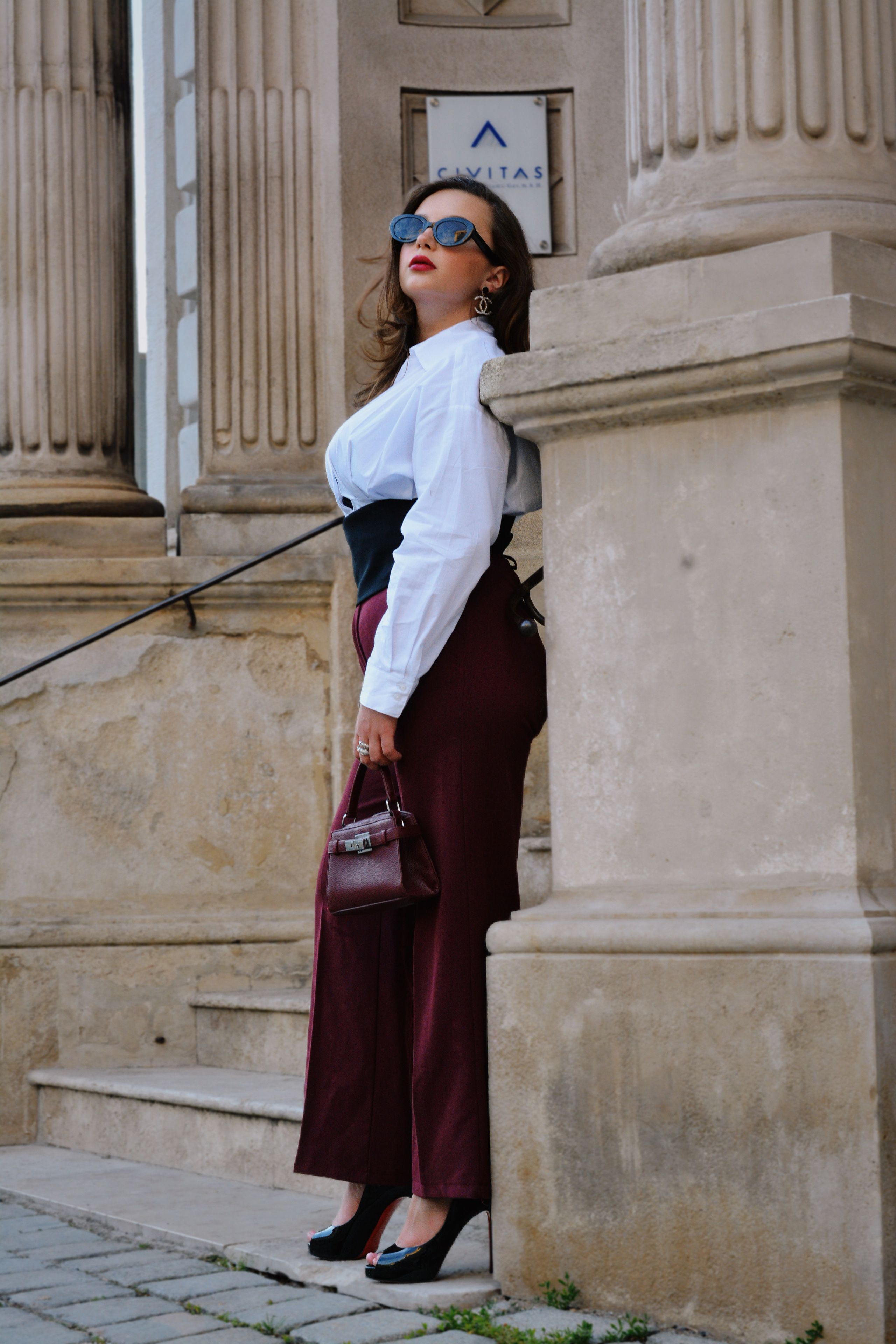A stylish woman in a white shirt and burgundy trousers leans against a classical stone column in Vienna, exuding modern elegance.