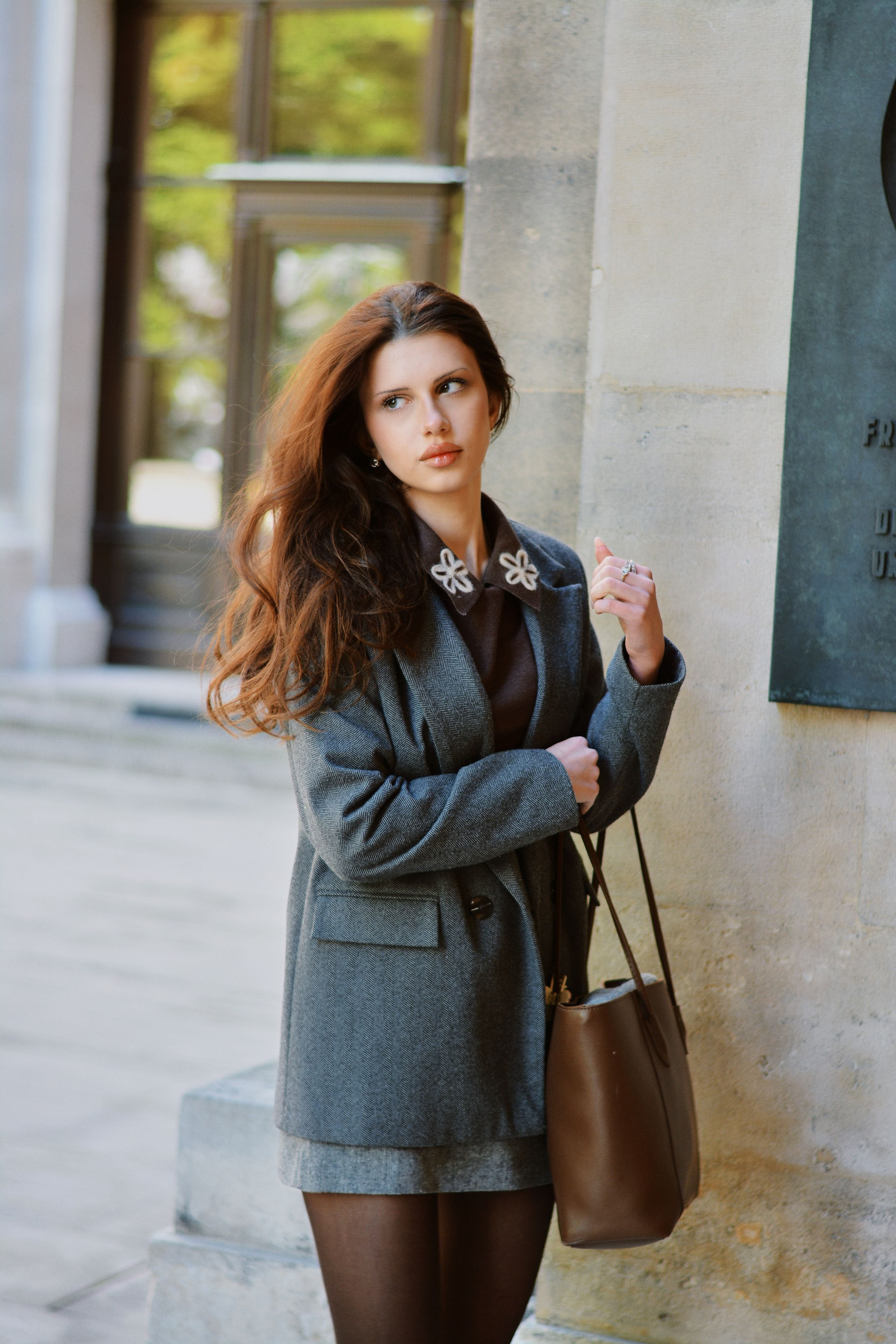 A stylish portrait of a young woman in a grey coat, standing against a classical stone facade in Vienna.