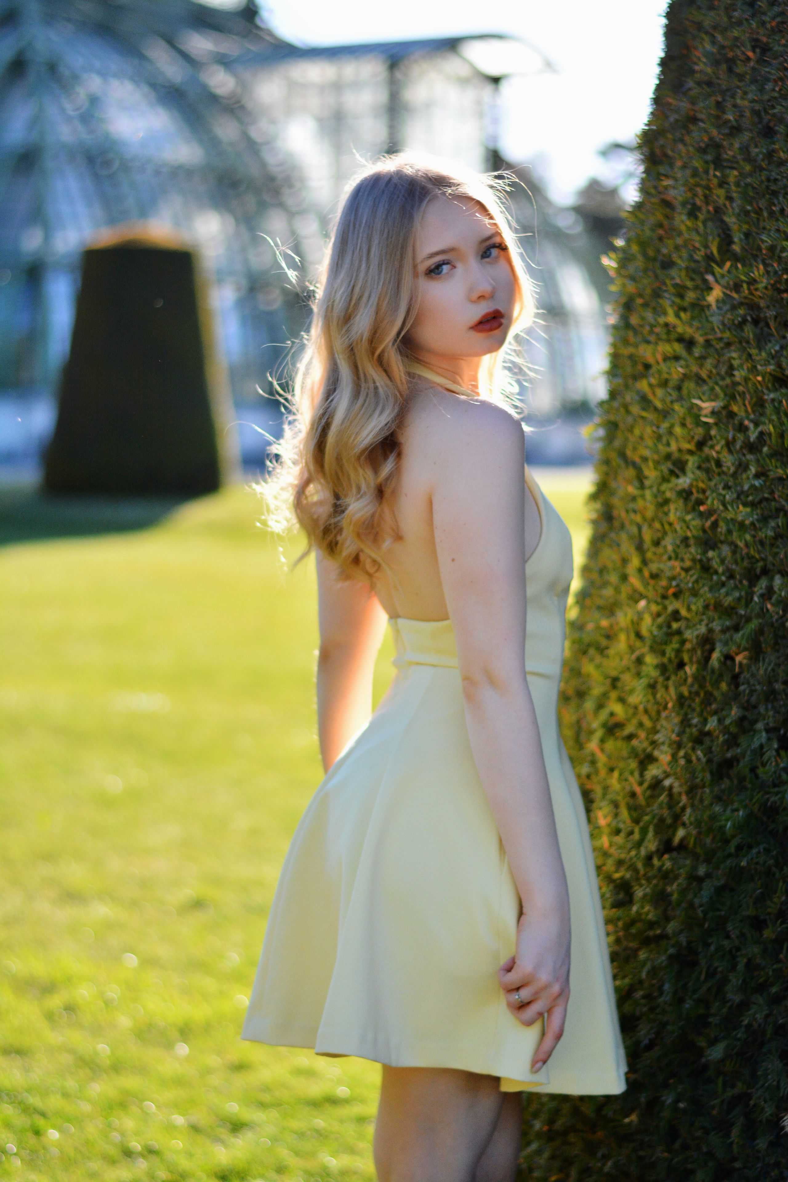 A beautiful young woman in a pale yellow dress poses outdoors in a lush green park setting, captured with soft natural light in Vienna.