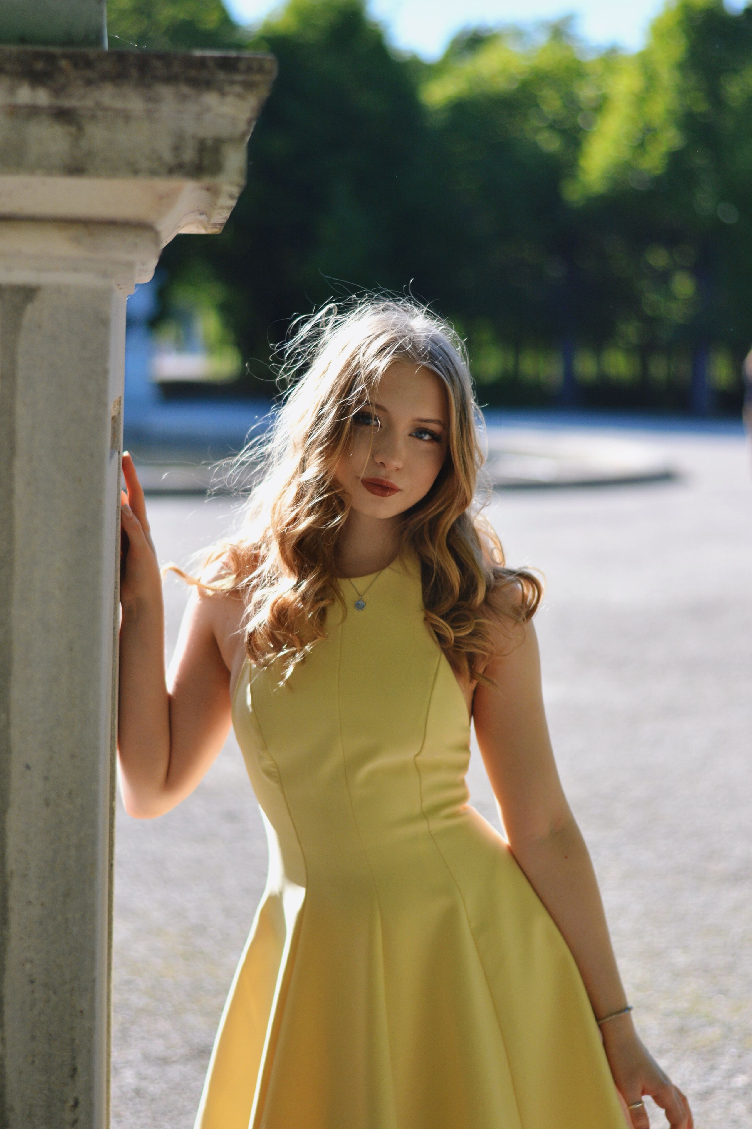 A beautiful young woman in a flowing yellow dress leans against a classical column in a lush Vienna park setting.