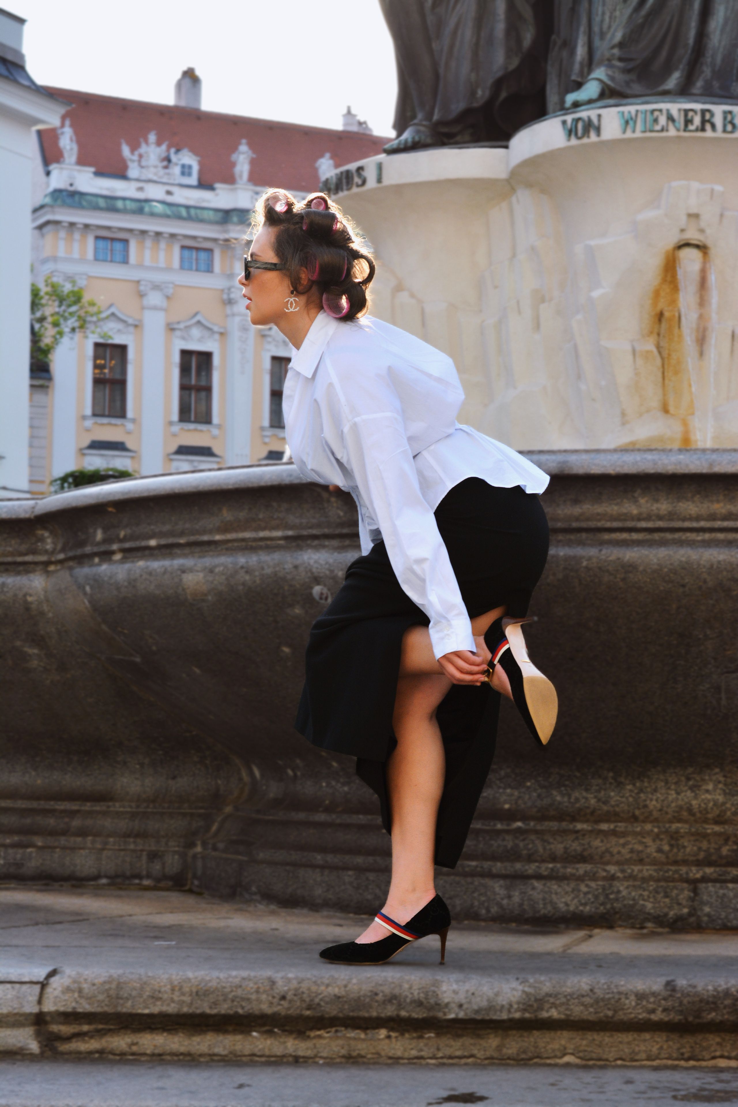 A stylish woman in a white shirt and black skirt poses near a grand statue in Vienna, capturing a modern portrait against classical architecture.