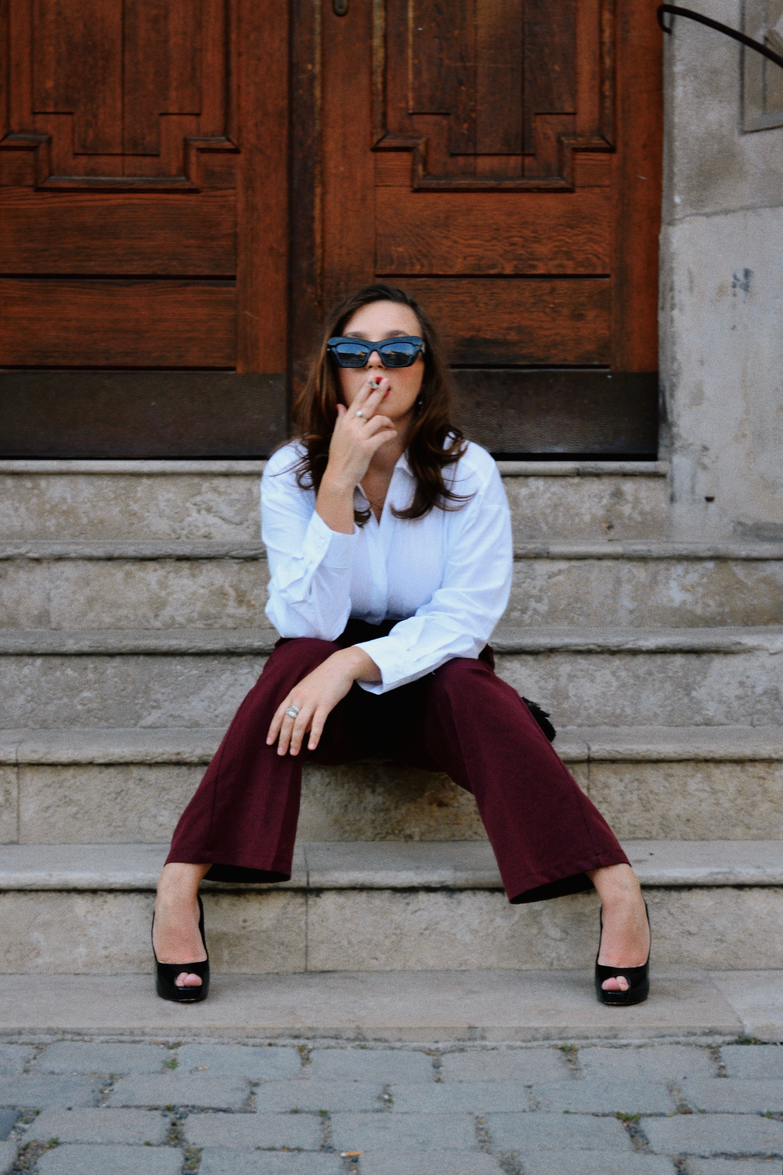 A stylish woman in a white shirt and burgundy trousers poses on stone steps outside a grand, dark wooden door in Vienna.