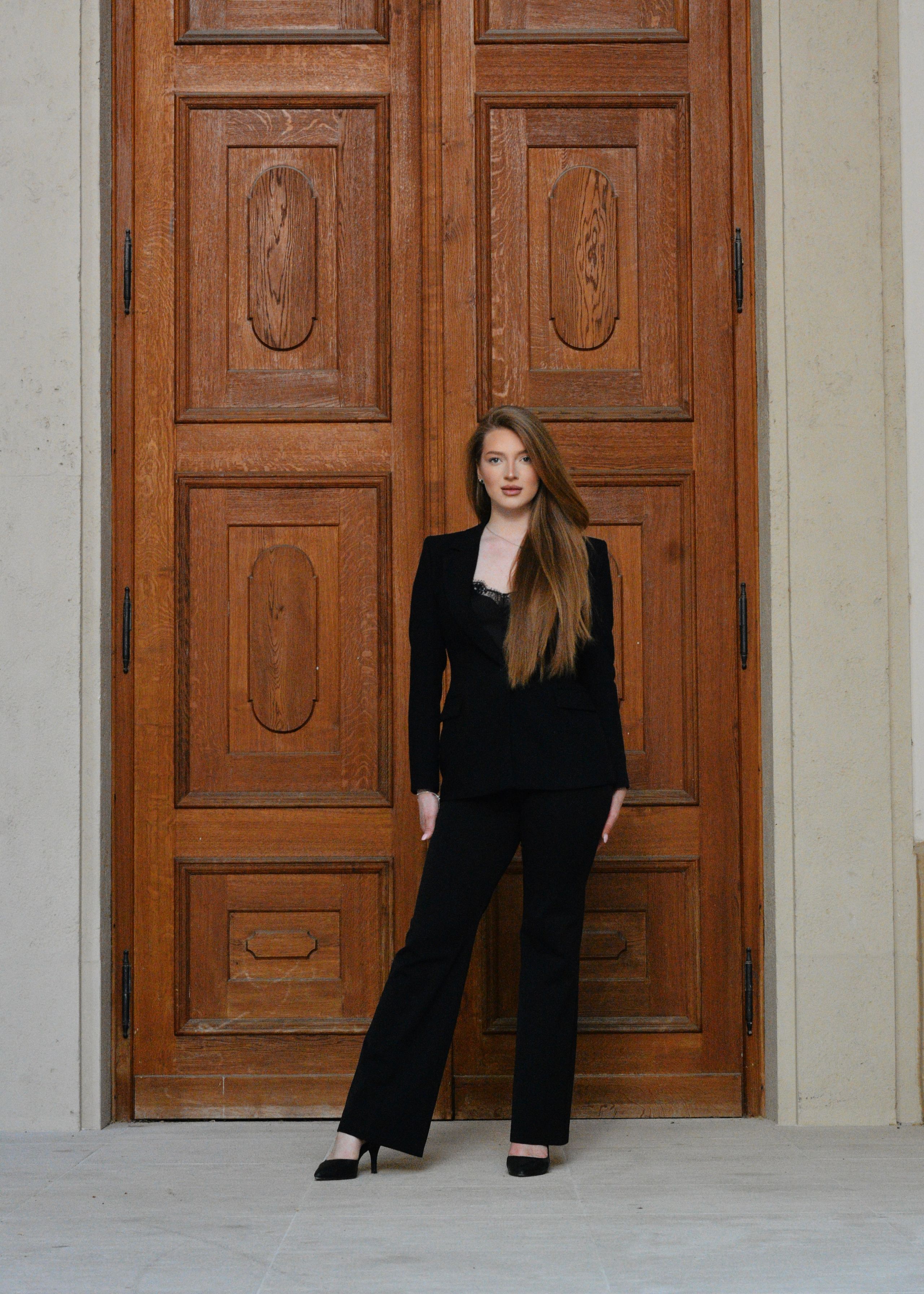 A stylish portrait of a young woman in a black suit standing against a grand, rustic wooden door in Vienna.
