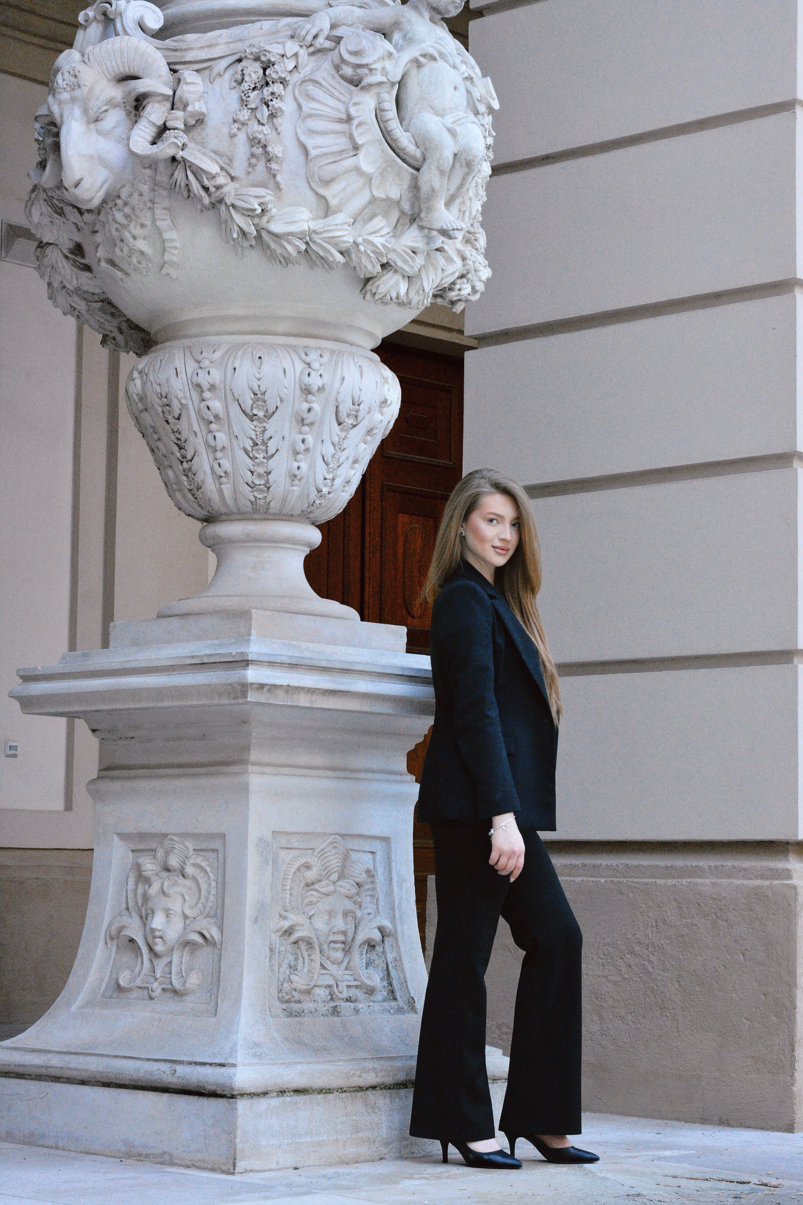 Elegant portrait of a woman in a black coat standing next to a classical stone urn in Vienna.