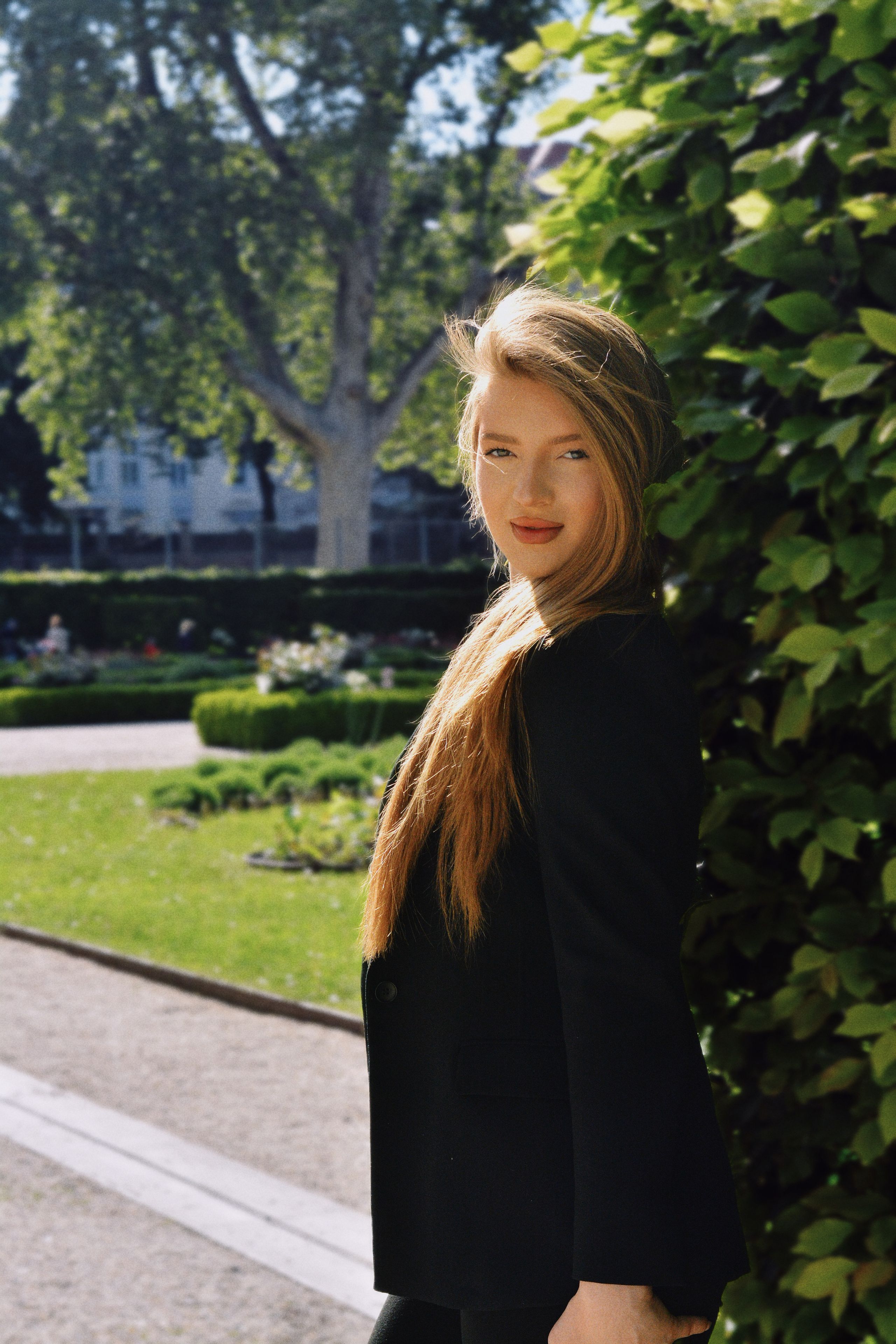 A beautiful portrait of a young woman with long blonde hair, standing outdoors in a lush, green Viennese park setting.