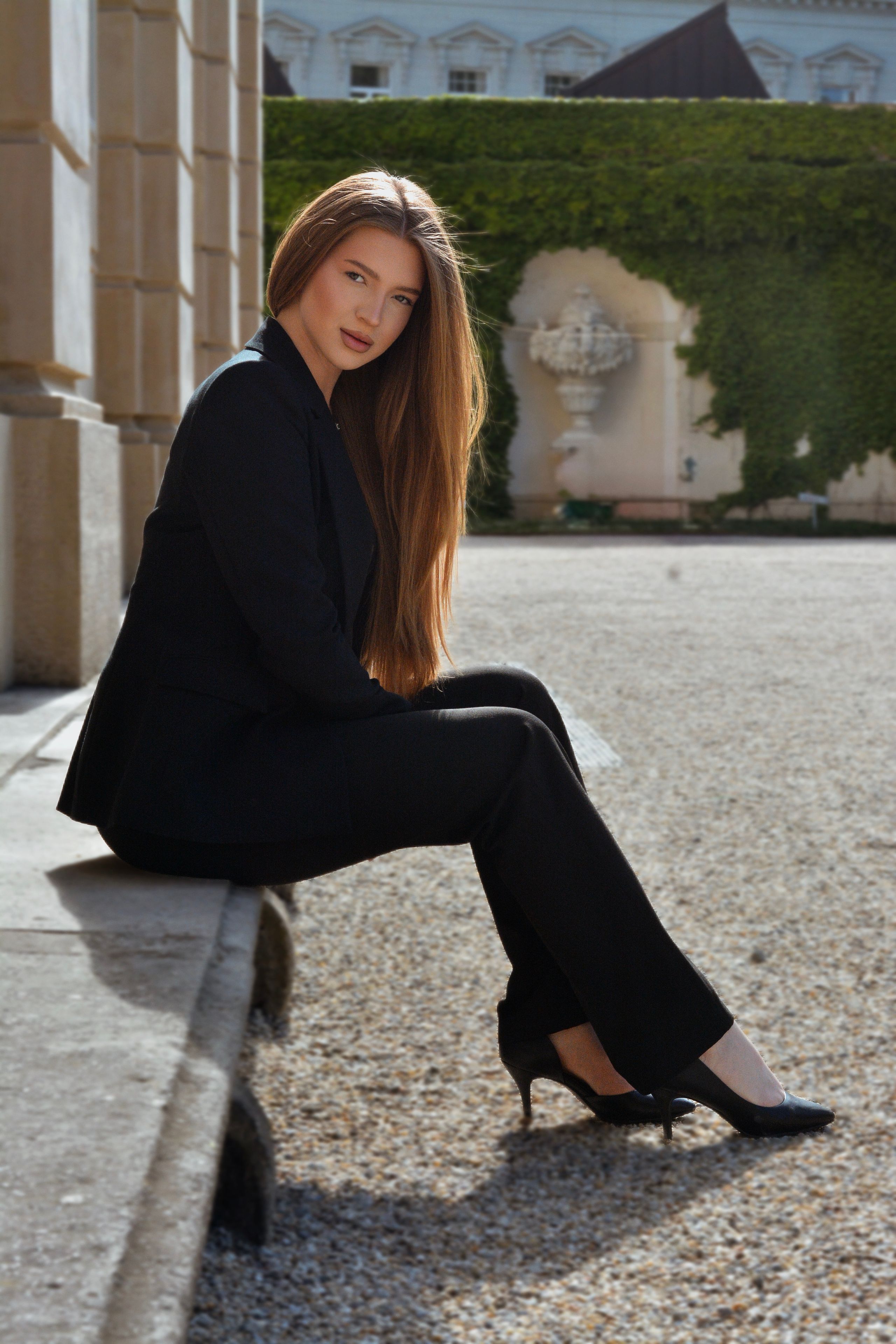 A striking portrait of a young woman in a black suit, seated outdoors against a formal, green-clad Viennese architectural backdrop.