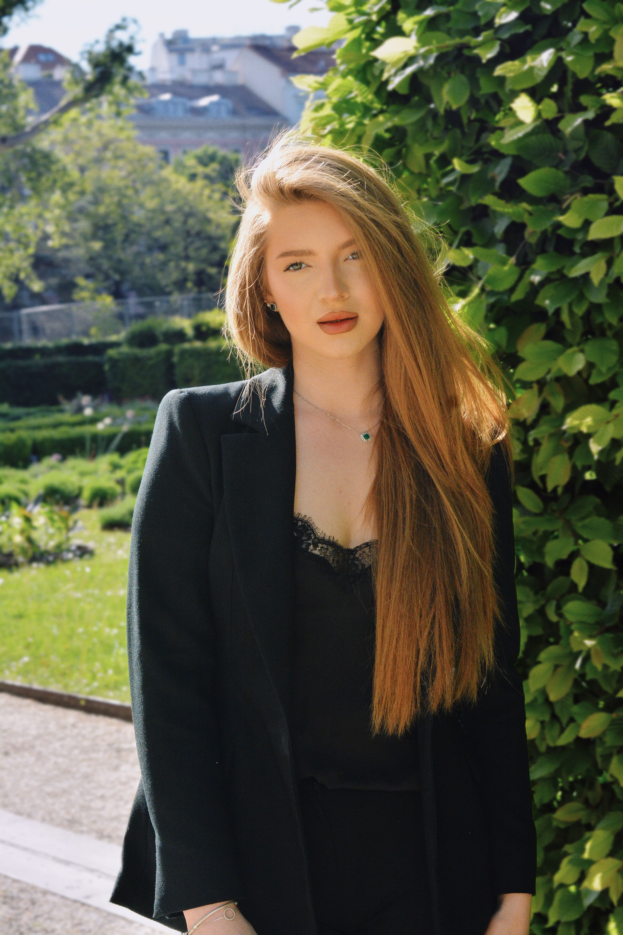 A striking portrait of a young woman with long blonde hair, set against lush greenery in a beautiful Vienna garden.