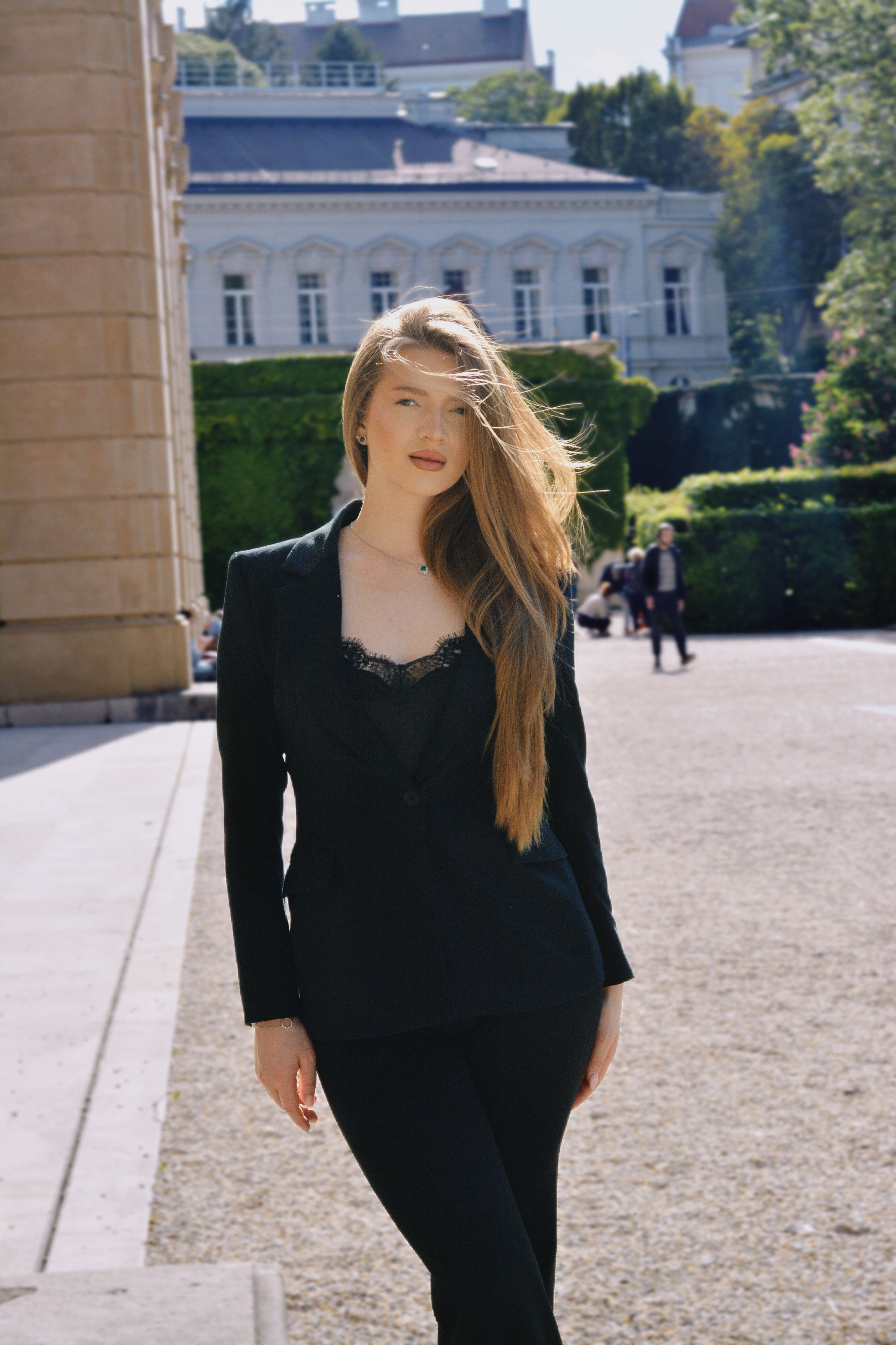 A striking portrait of a young woman in a black blazer, standing outdoors in a grand, classical Vienna setting.
