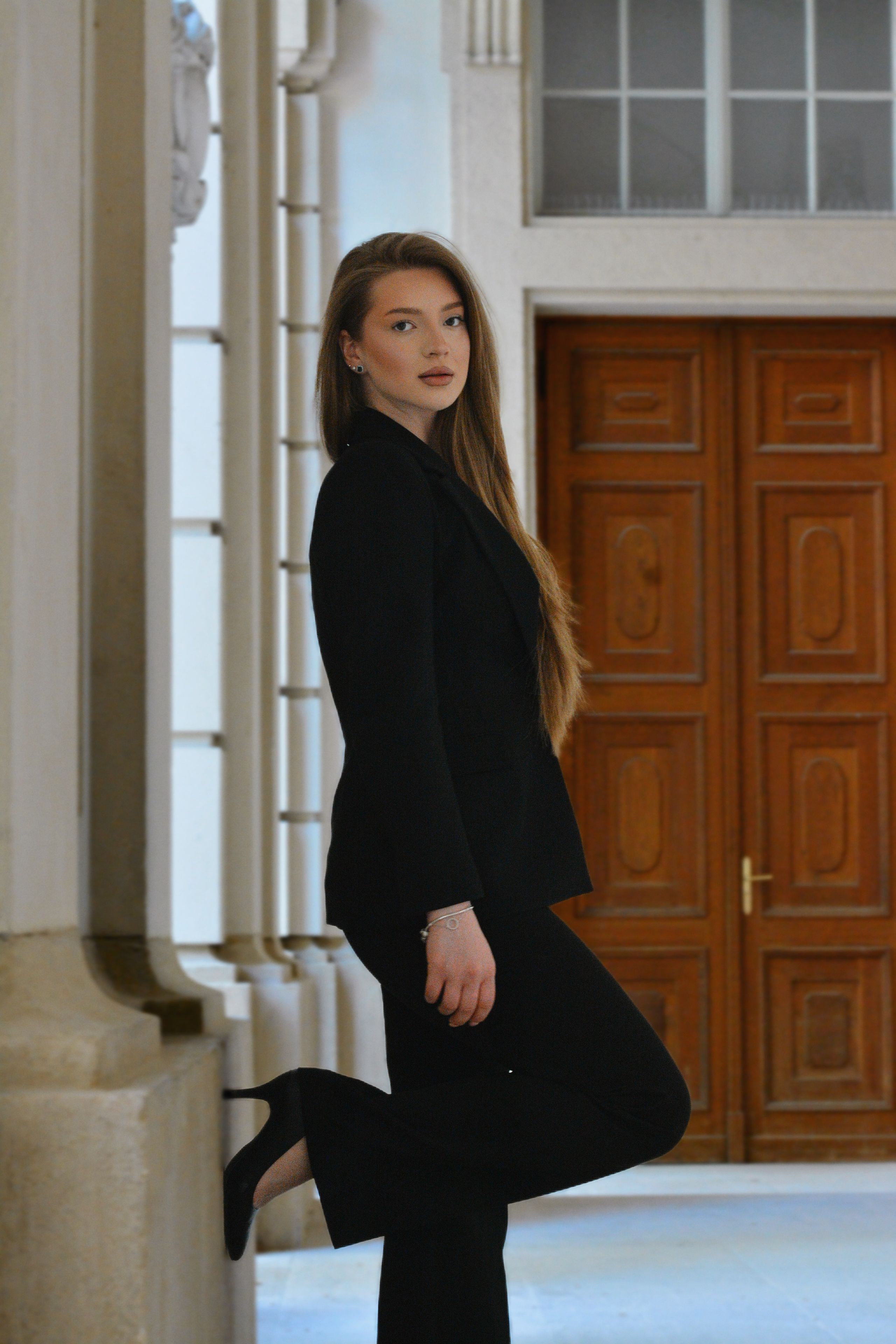 Elegant portrait of a young woman in a black suit, standing in a grand, classical Viennese architectural hallway.